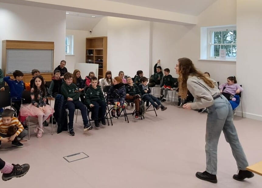 Fiona is standing in front of a group of children, some of whom have their hands raised. She is leaning forward and smiling.
