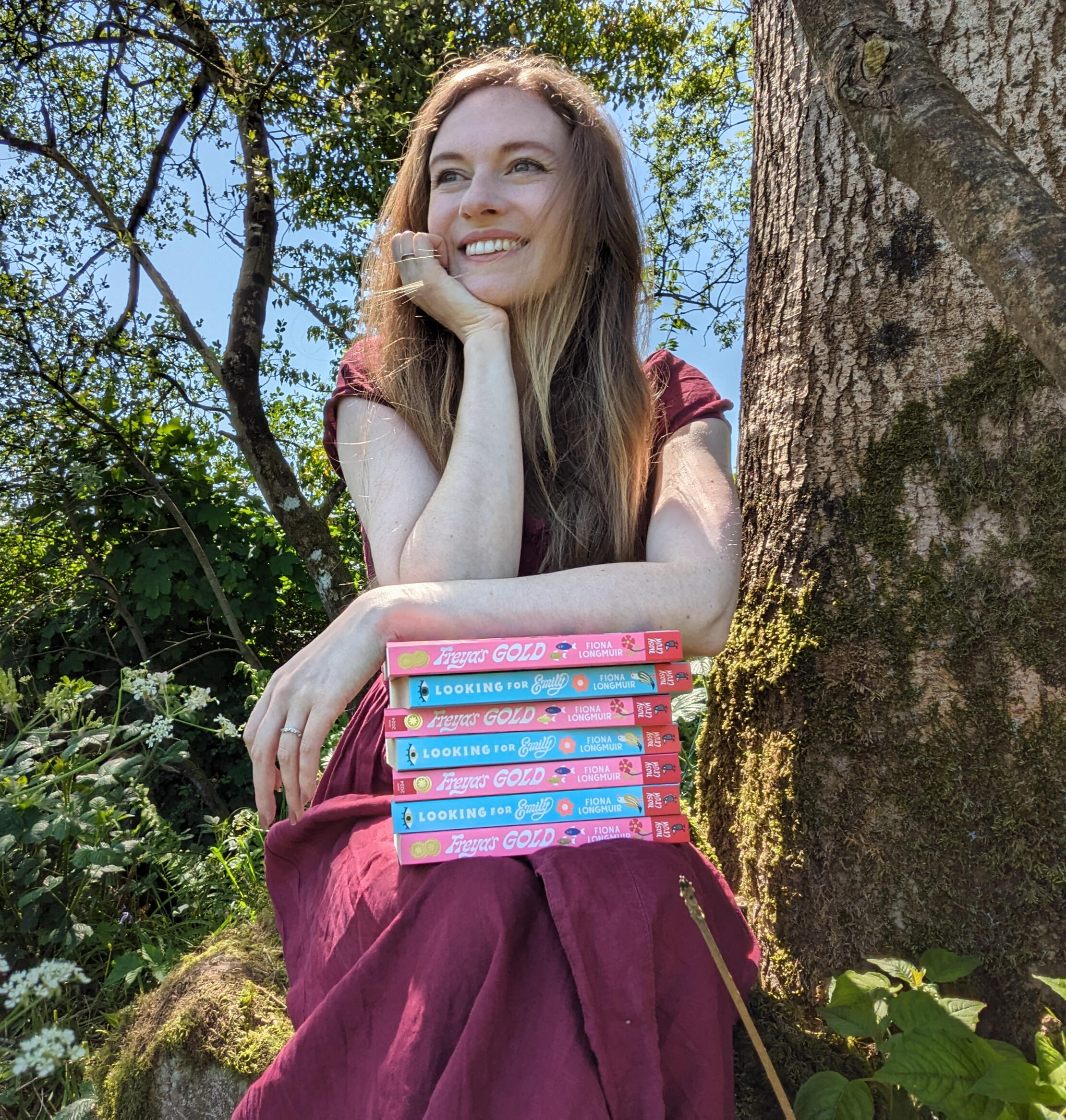 Fiona is looking to the side of the camera and laughing. She is holding a stack of Looking for Emily and Freya's Gold books and wearing a burgundy linen dress. She is sitting in front of a tree surrounded by frothy white cow parsley.